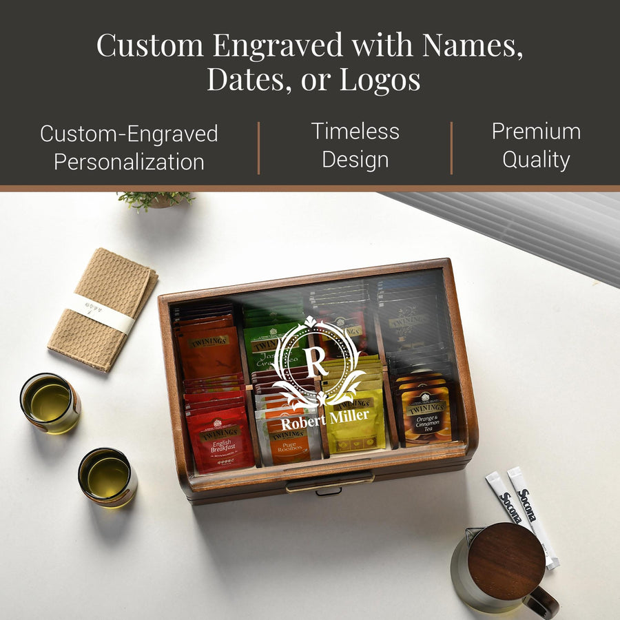 a wooden box with a glass front, containing various tea bags and a small plant. The box has a personalized engraving of a name, and there are two cups and a small plant on the table.