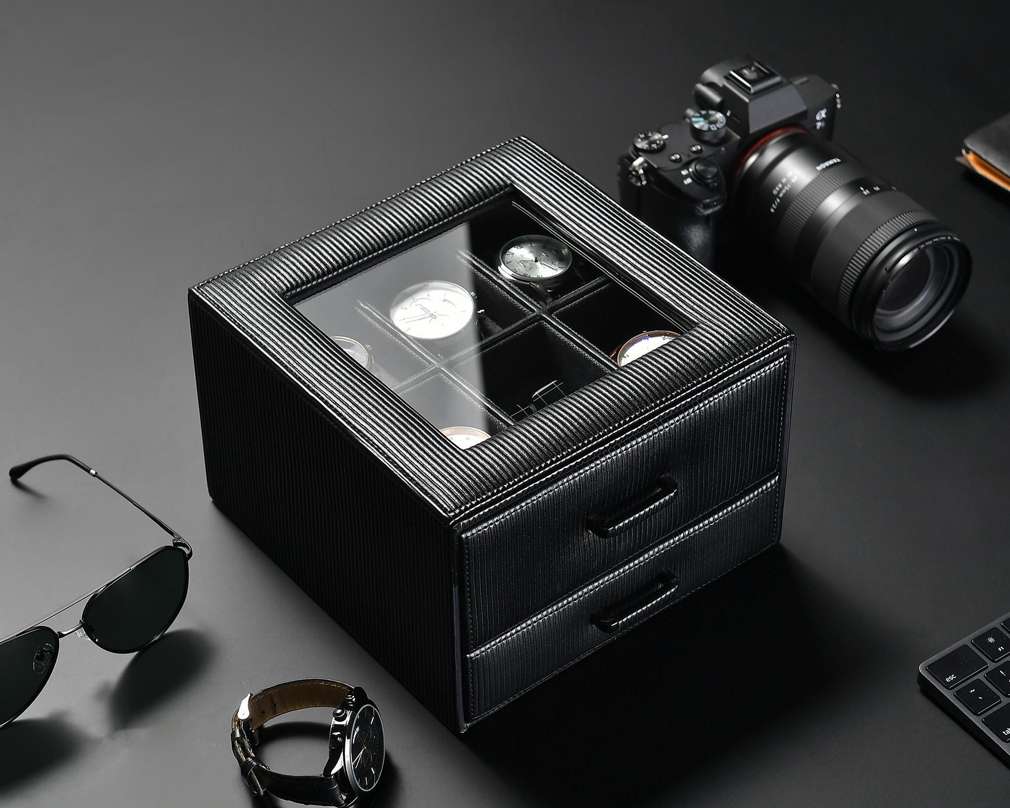 A black watch box with a glass top displaying six watches is placed on a dark surface. Below the display are two pull-out drawers. Surrounding the box are a camera with a lens, a pair of sunglasses, a wristwatch, and a keyboard corner.
