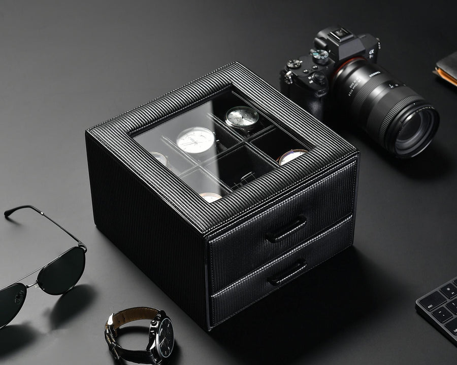 A black watch box with a glass top displaying six watches is placed on a dark surface. Below the display are two pull-out drawers. Surrounding the box are a camera with a lens, a pair of sunglasses, a wristwatch, and a keyboard corner.