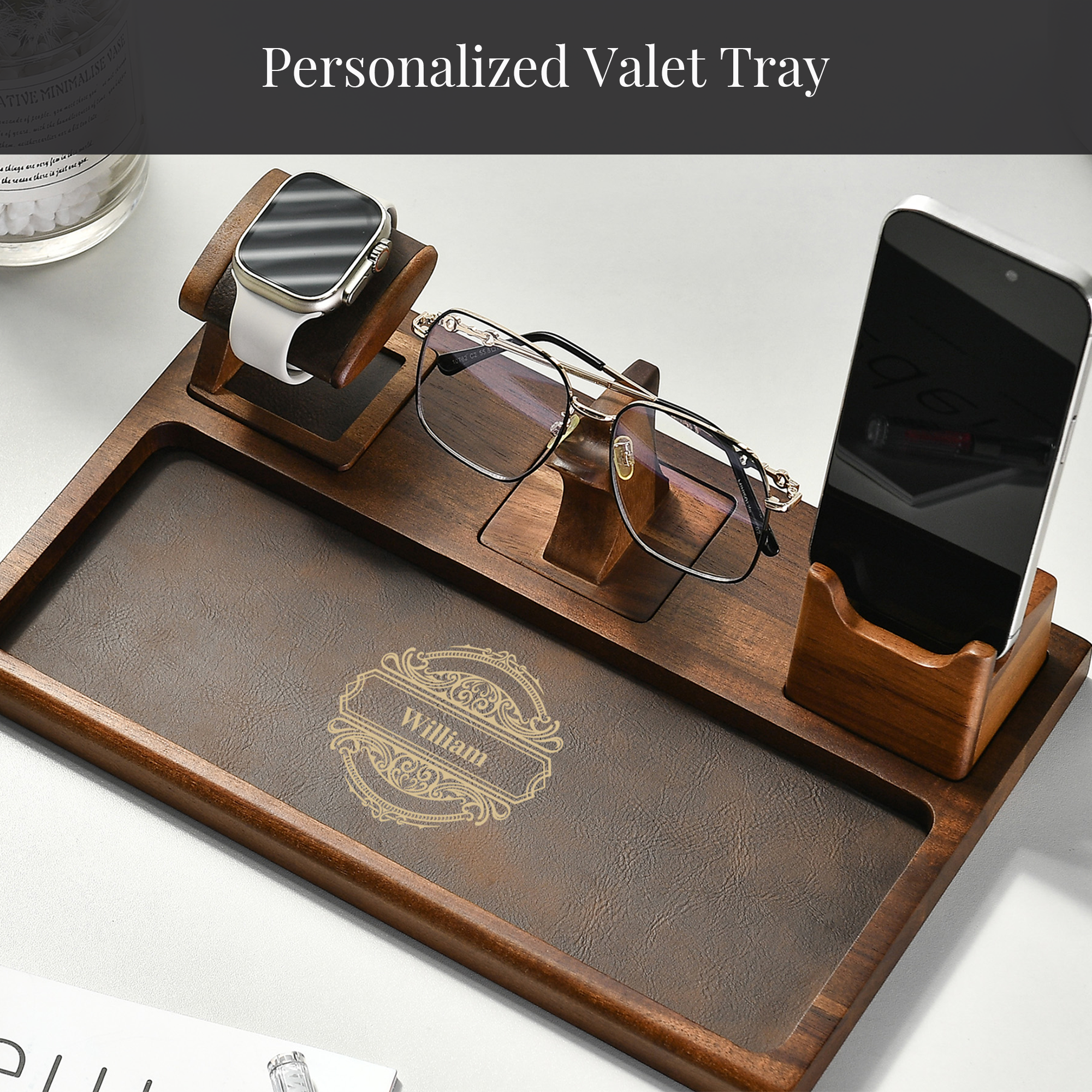 A wooden tray with a smartphone, glasses, and a watch on it, along with a personalized name engraved on the tray.