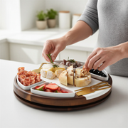 A person is arranging a variety of food items on a wooden platter, which includes cheese, bread, fruit, and other appetizers.