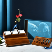a wooden serving tray with three white plates, a wooden box with a blue design, a bottle of wine, a vase with red roses, and a wooden tray with various utensils.