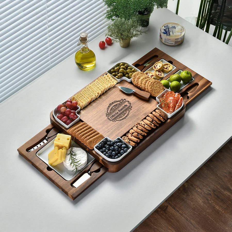 A wooden serving platter with various food items, including crackers, cheese, grapes, and olives, arranged on a white table.