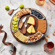 A wooden platter filled with an assortment of cheese, crackers, nuts, and fruit, accompanied by a bottle of whiskey and a glass of wine.