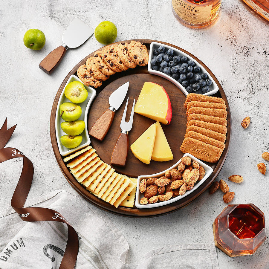A wooden platter filled with an assortment of cheese, crackers, nuts, and fruit, accompanied by a bottle of whiskey and a glass of wine.