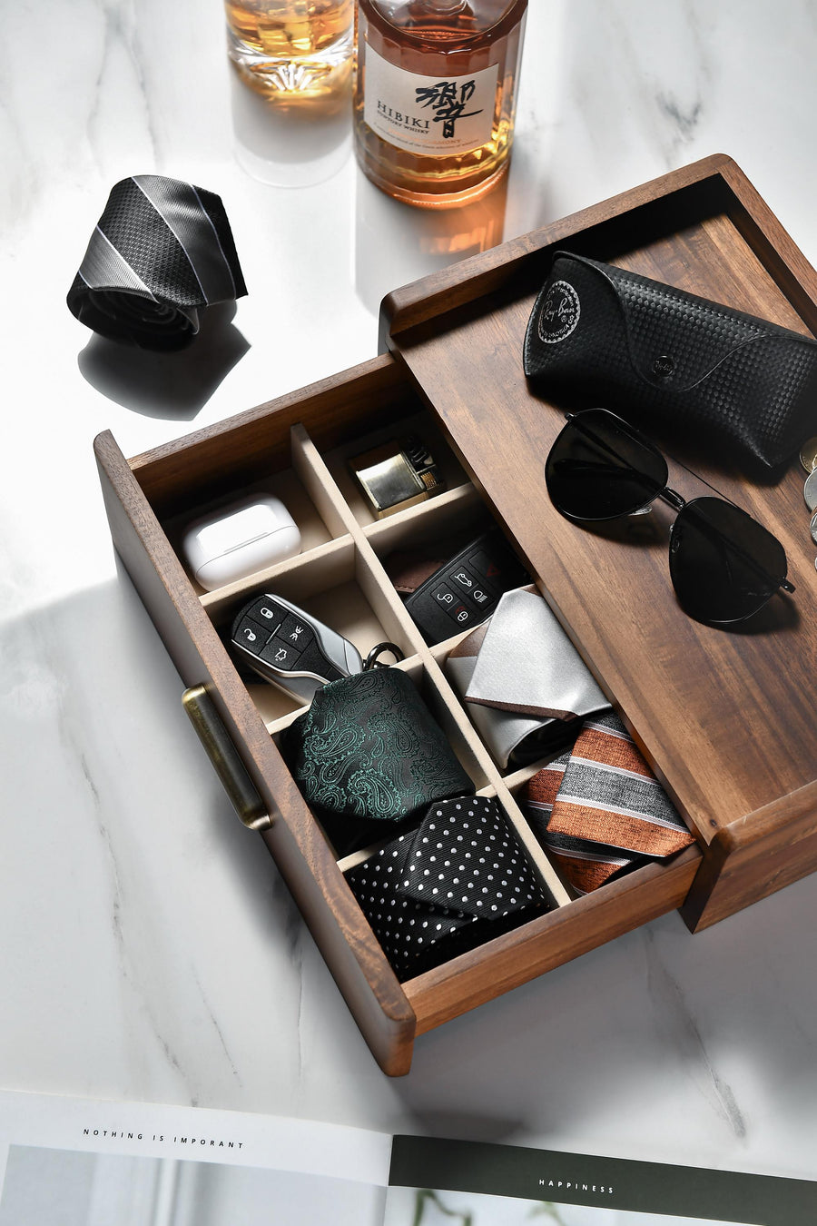 a wooden drawer with compartments, containing various items such as a tie, sunglasses, a phone, and other accessories.