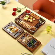 a wooden tray filled with various snacks and appetizers, including crackers, cheese, nuts, and fruits, arranged on a table. There are also some flowers and a book visible in the scene.