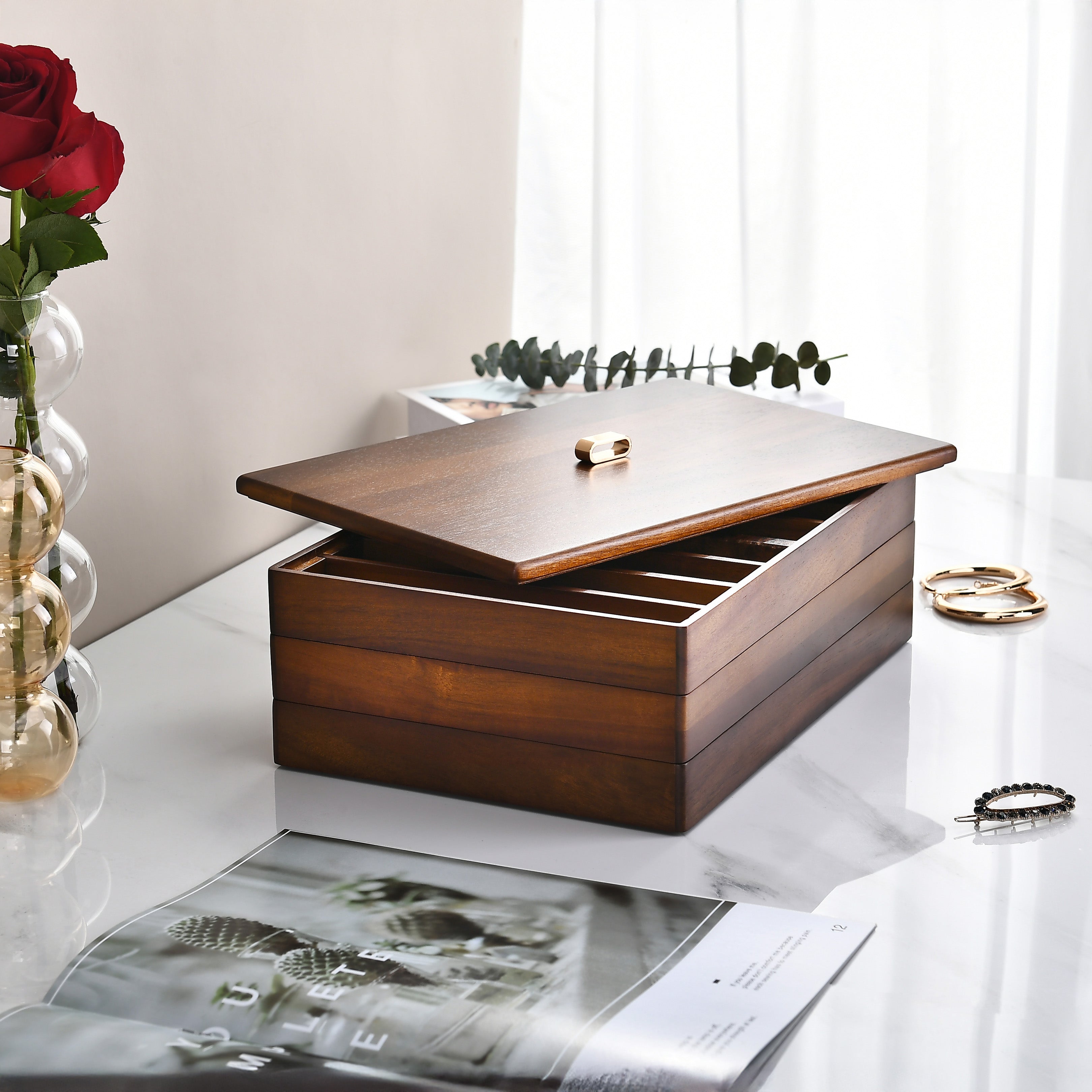 a wooden jewelry box with a lid, placed on a white marble countertop. The box has a few gold rings visible inside. Next to the box, there is a vase with a red rose, and a magazine or book is also present on the countertop.