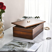 a wooden jewelry box with a lid, placed on a white marble countertop. The box has a few gold rings visible inside. Next to the box, there is a vase with a red rose, and a magazine or book is also present on the countertop.