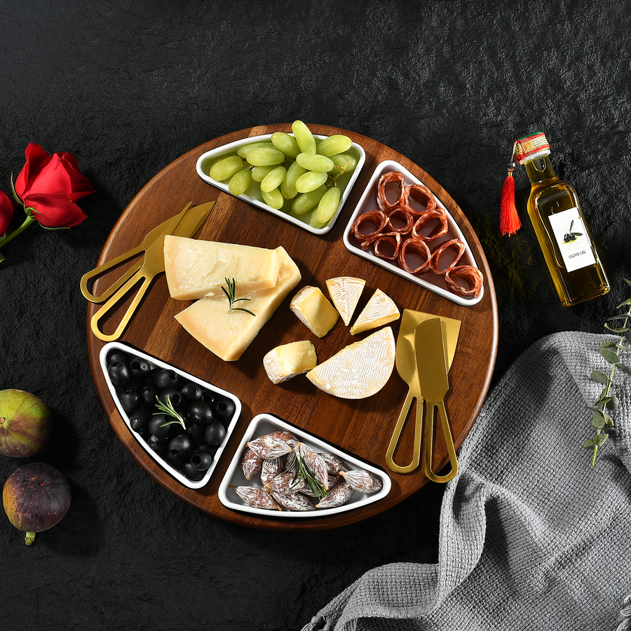 A wooden platter with various cheese slices, grapes, olives, and other snacks arranged on a dark surface.