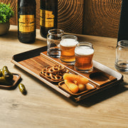 A wooden tray with three glasses of beer, a plate of pretzels, and a small dish of pickles, accompanied by two bottles of beer, one of which is Budweiser.