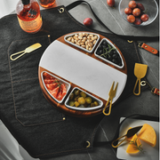 a round wooden platter with various food items, including olives, cheese, and nuts, arranged on a table. There are also some bottles and a bowl of fruit present in the scene.