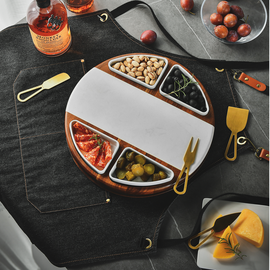 a round wooden platter with various food items, including olives, cheese, and nuts, arranged on a table. There are also some bottles and a bowl of fruit present in the scene.