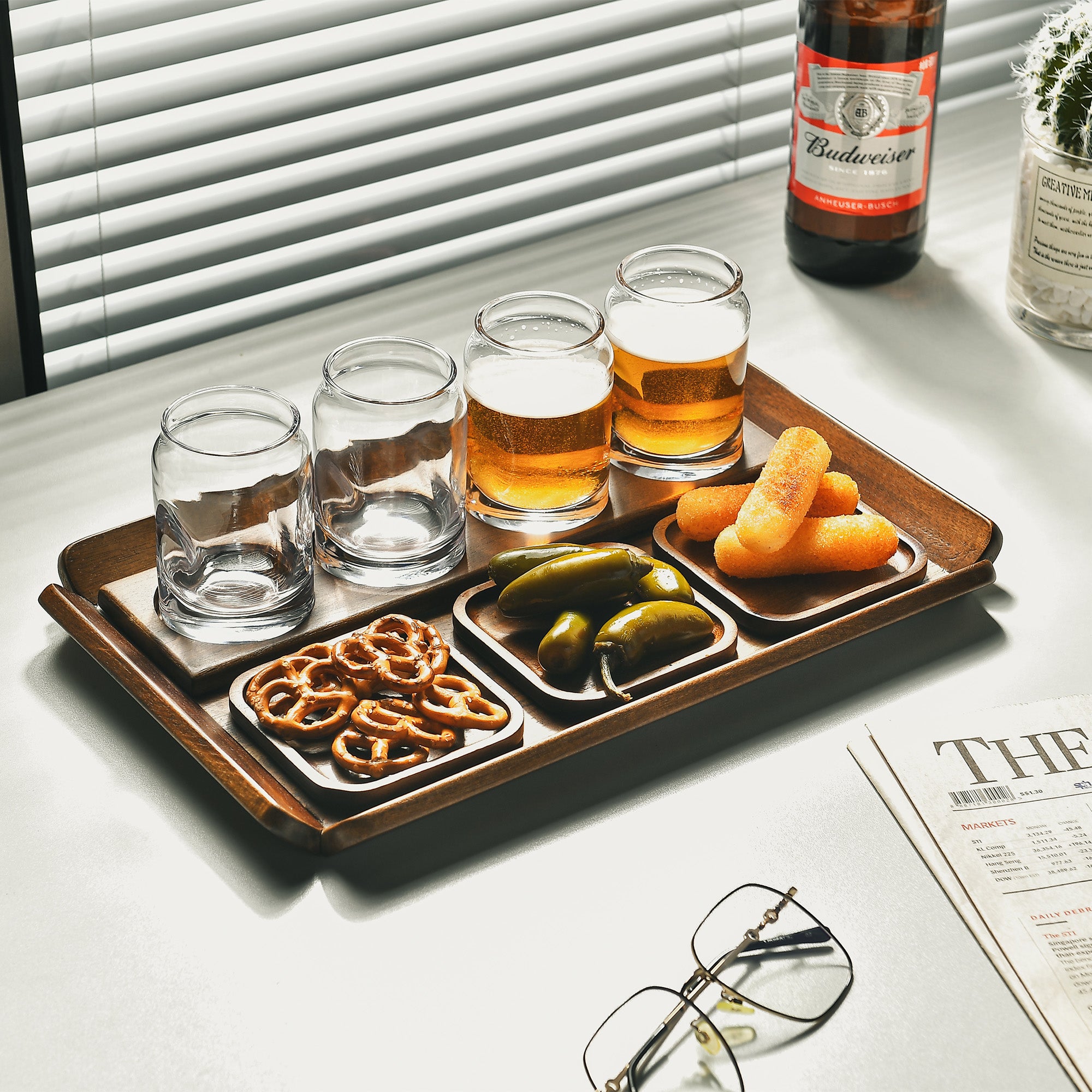A tray with four glasses of beer, a plate of pickles, and a bottle of Budweiser beer is displayed on a table.