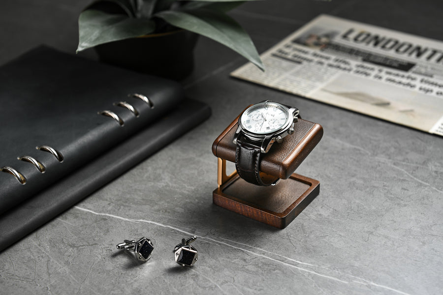 A wooden watch stand with a silver watch and two silver cufflinks on a gray surface, with a black notebook and a newspaper in the background.