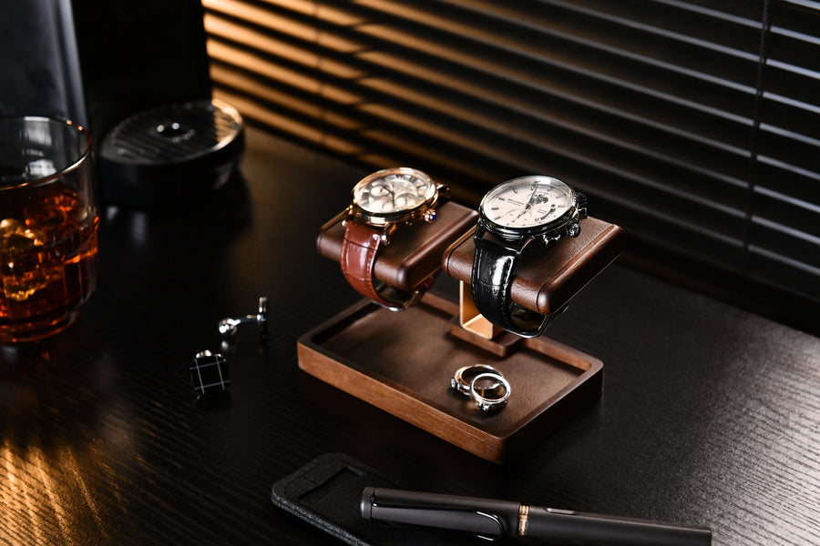 a collection of luxury items, including two wristwatches, a pen, a glass of amber liquid, and a wooden tray with rings and a pen.