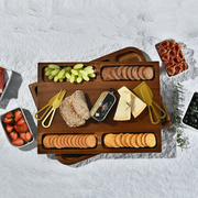 A wooden tray filled with various food items, including grapes, bread, cheese, crackers, and other snacks, arranged on a white surface.