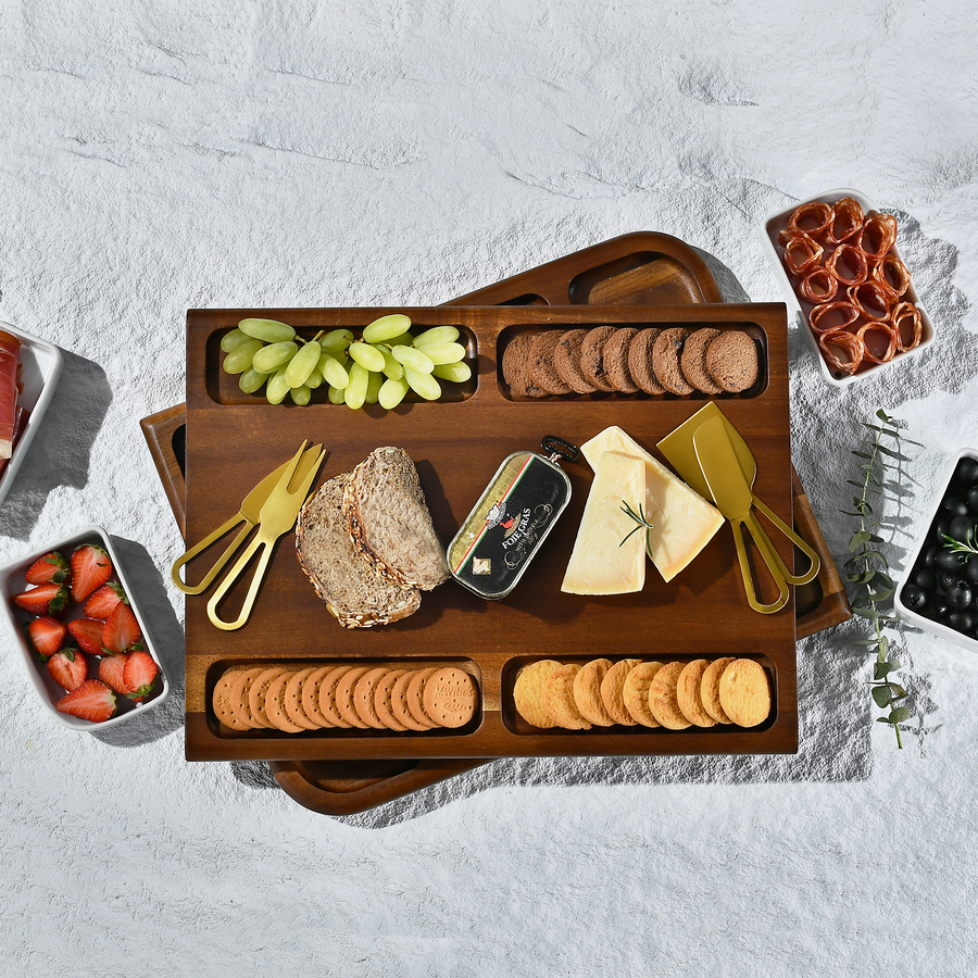 A wooden tray filled with various food items, including grapes, bread, cheese, crackers, and other snacks, arranged on a white surface.
