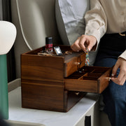 A person is sitting on a couch, opening a wooden jewelry box with multiple compartments.