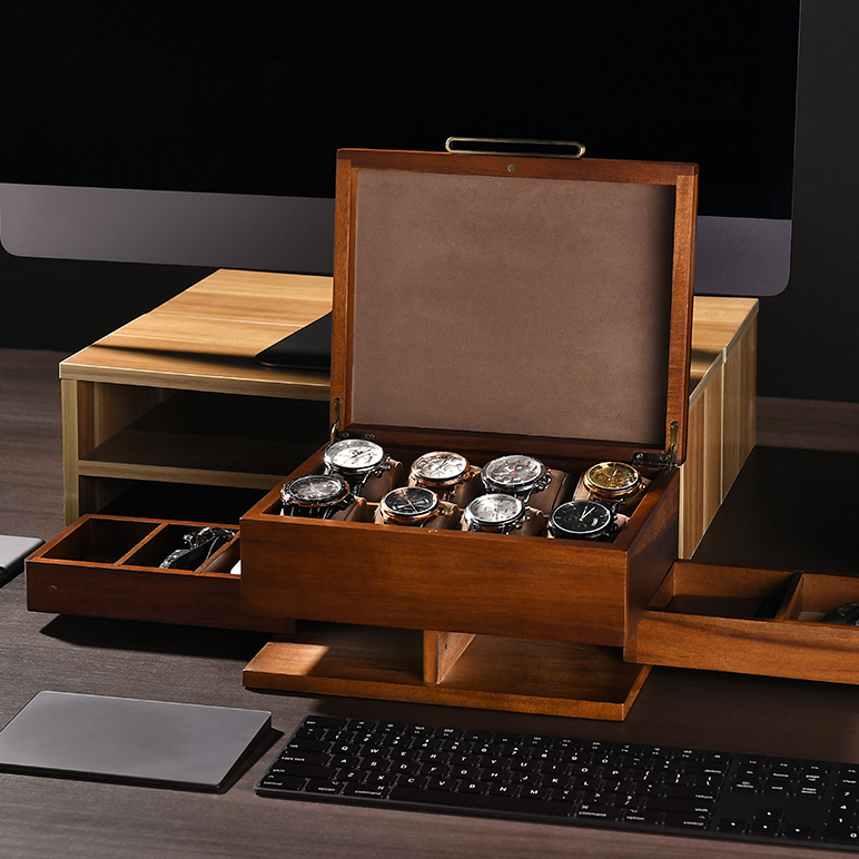 A wooden watch box containing multiple watches is placed on a desk, with a computer keyboard and other office supplies visible in the background.