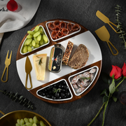 a round wooden platter with various food items arranged on it, including grapes, cheese, bread, olives, and other snacks. The platter is placed on a dark surface, and there are also some decorative elements like a rose and a knife visible in the background.