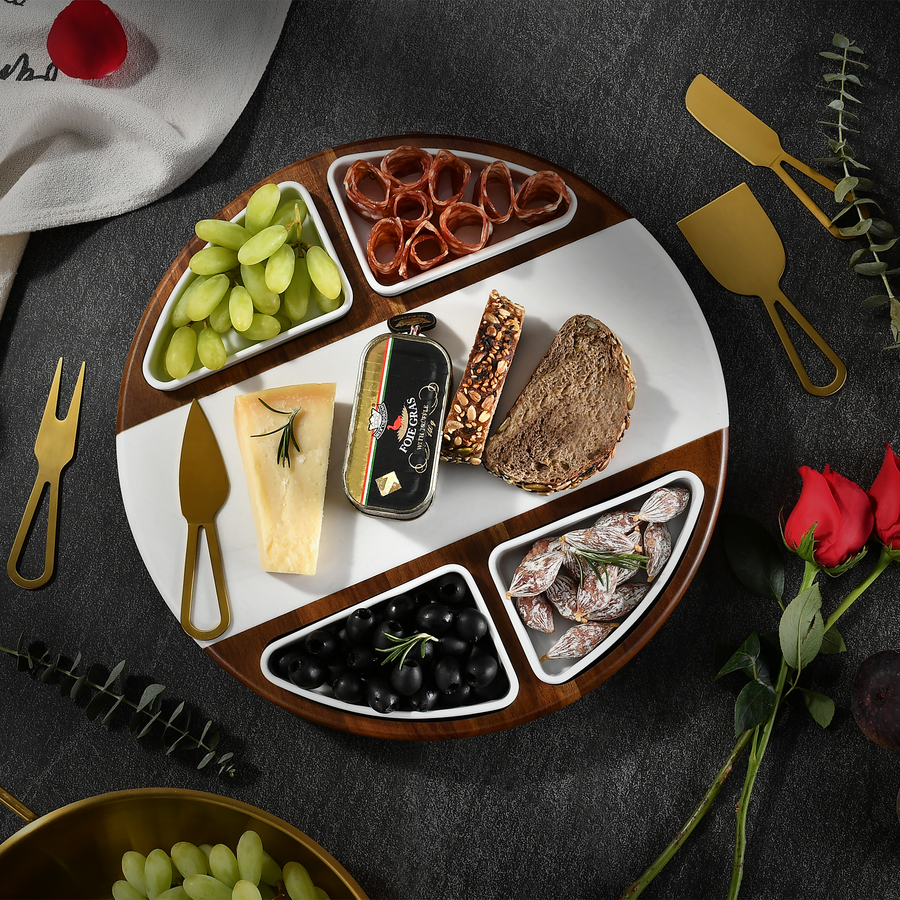 a round wooden platter with various food items arranged on it, including grapes, cheese, bread, olives, and other snacks. The platter is placed on a dark surface, and there are also some decorative elements like a rose and a knife visible in the background.