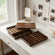a wooden jewelry box with multiple compartments, placed on a white desk. The box has a wooden lid and is open, revealing the contents inside. The desk is positioned near a window, and there is a mirror and a framed photo on the wall behind the jewelry box.