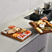 A person is holding a wooden tray filled with various appetizers and snacks, including crackers, bread, cheese, and fruit, in a kitchen setting.