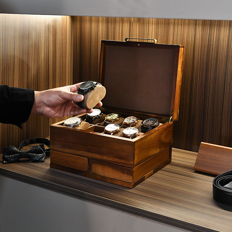 A person's hand is holding a watch over a wooden box containing several watches, which is placed on a wooden surface.