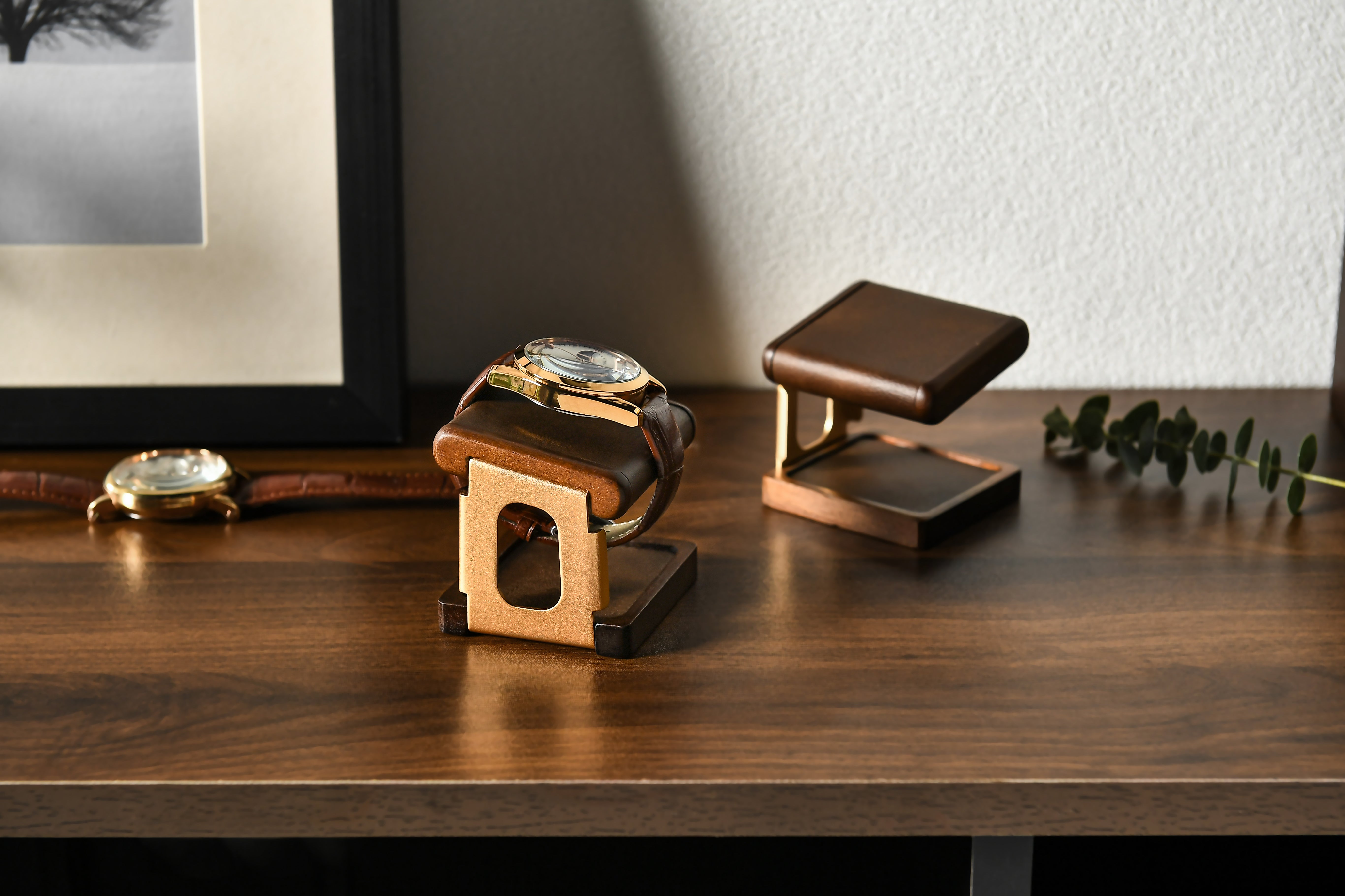 a wooden stand with two watches placed on it, one of which is a gold watch. The stand is positioned on a wooden surface, and there is a framed picture in the background.