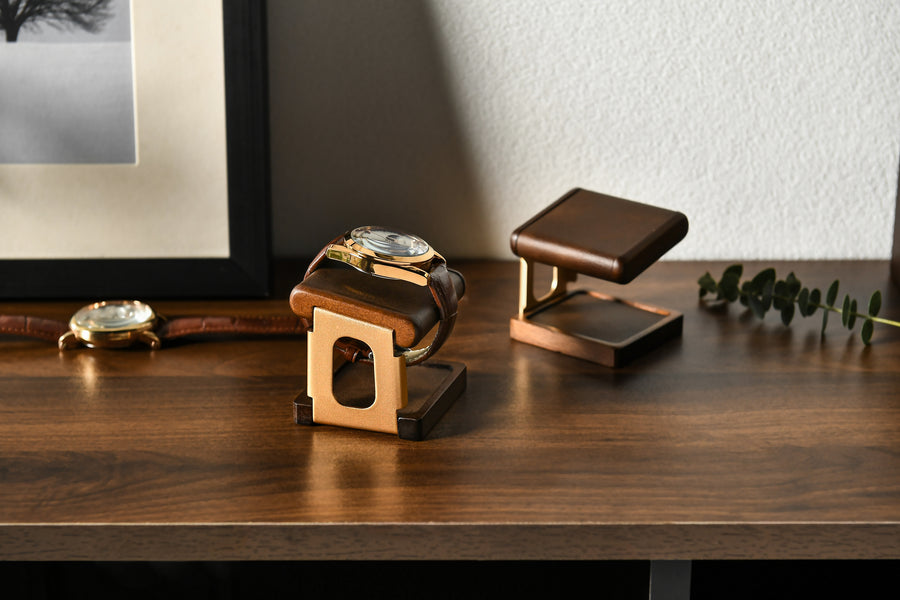 a wooden stand with two watches placed on it, one of which is a gold watch. The stand is positioned on a wooden surface, and there is a framed picture in the background.