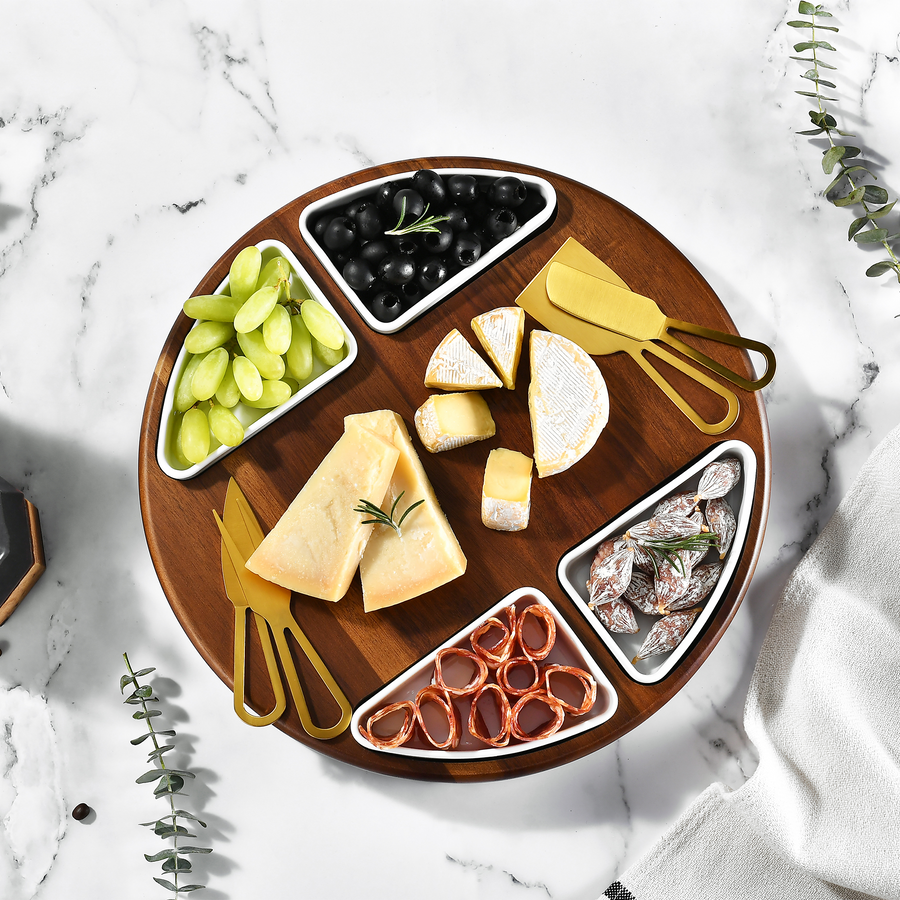 A wooden platter with various cheese slices, grapes, olives, and other appetizers arranged on a marble surface.