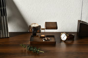 A wooden stand displaying three watches and a ring on a wooden tray, with a plant and a black object in the foreground.