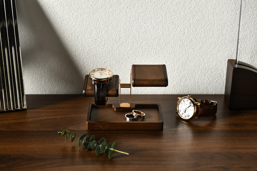 A wooden stand displaying three watches and a ring on a wooden tray, with a plant and a black object in the foreground.