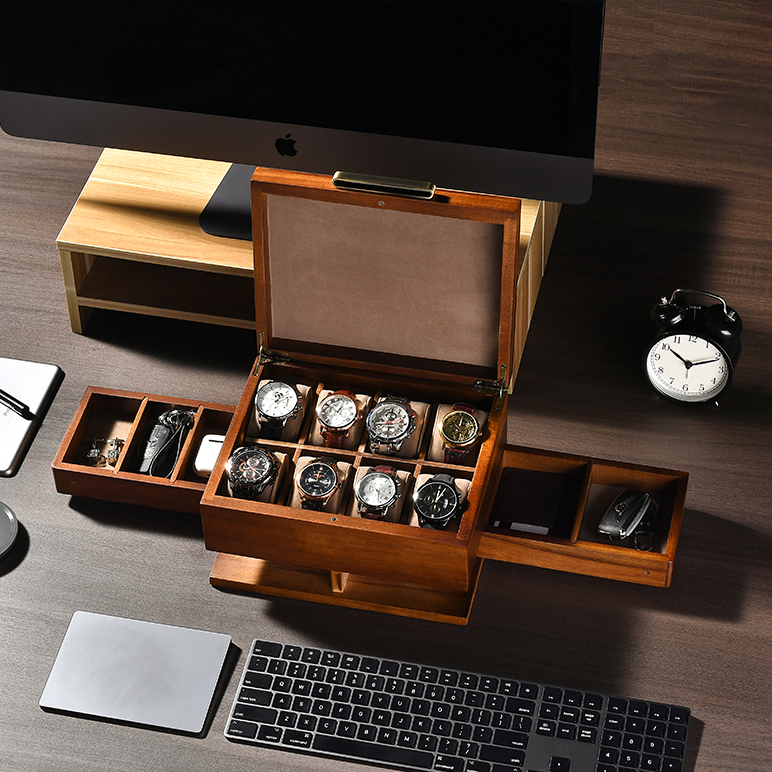 a wooden watch storage box with multiple compartments, containing various watches, placed on a desk alongside a computer keyboard, a clock, and other office supplies.