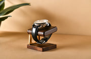 A black leather watch with a silver face is displayed on a wooden stand, placed on a beige background.