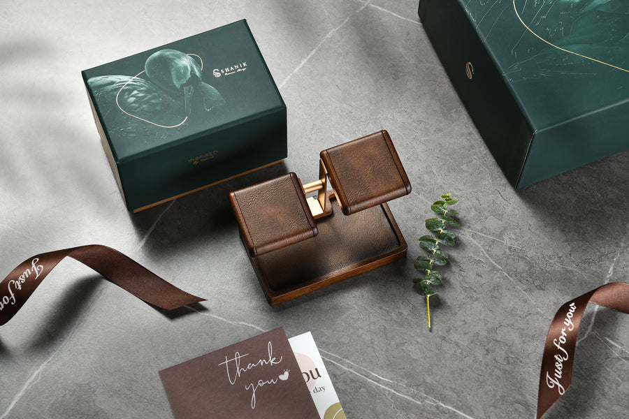 a set of three items arranged on a marble surface: a small wooden box with a picture of a bird on it, a brown leather box, and a brown ribbon. There is also a small card with a message that reads "Thank you".