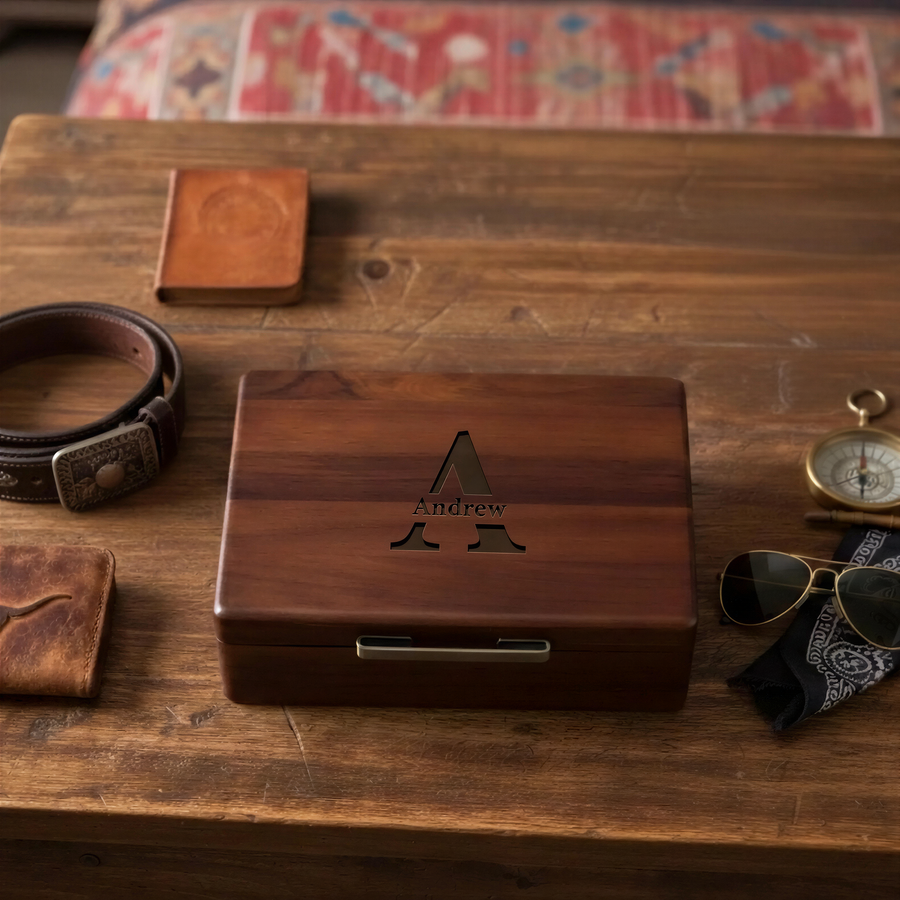 a wooden box with a personalized name carved into it, sitting on a wooden surface alongside various personal items such as a belt, sunglasses, and a wallet.