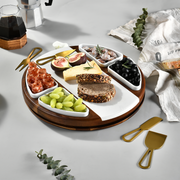 a wooden serving platter with various food items, including bread, cheese, grapes, figs, and olives, arranged on a white surface. There are also some gold serving utensils, such as a knife and a pair of tongs, placed next to the platter.