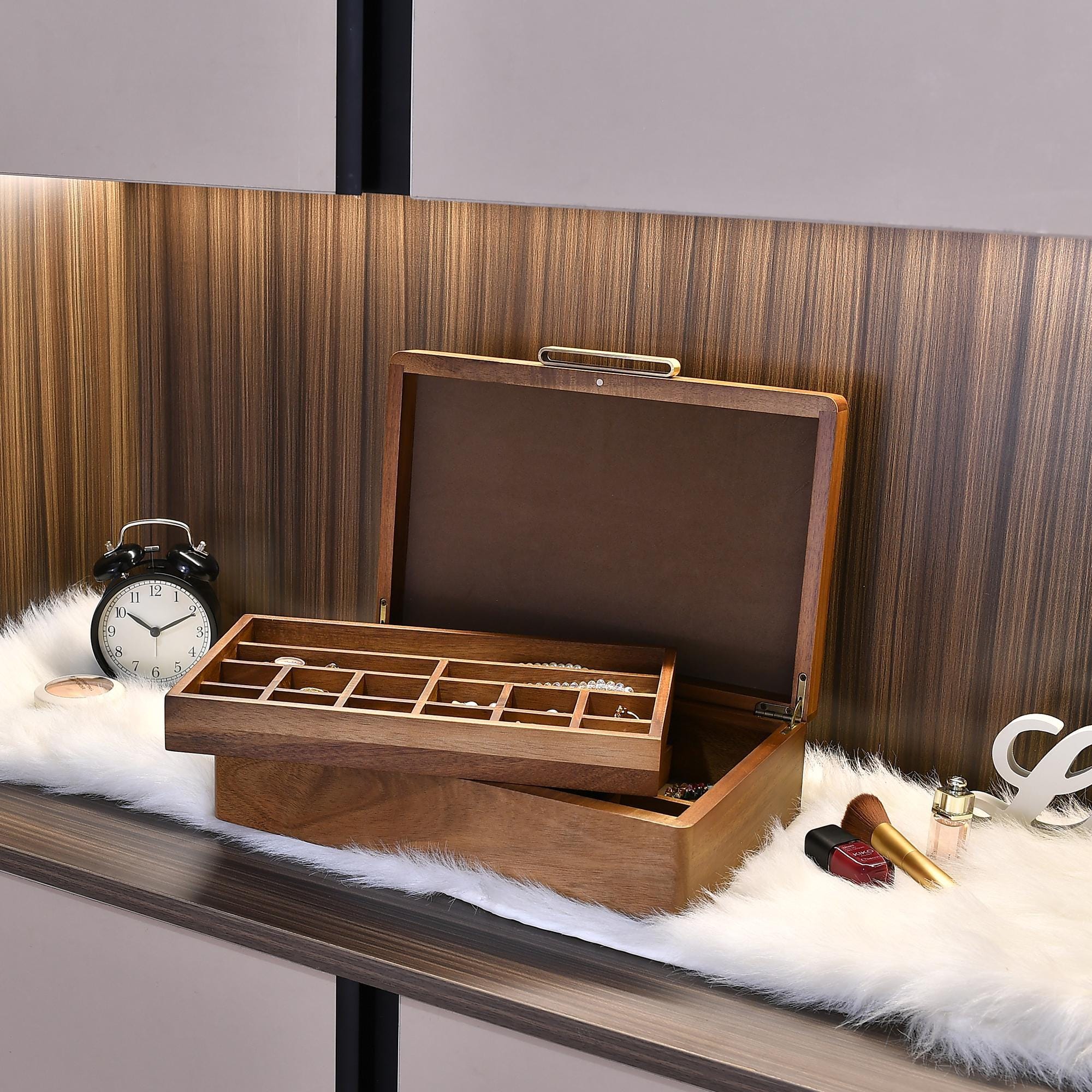 a wooden jewelry box with compartments, placed on a white fur rug. There is a black alarm clock and a makeup brush nearby.