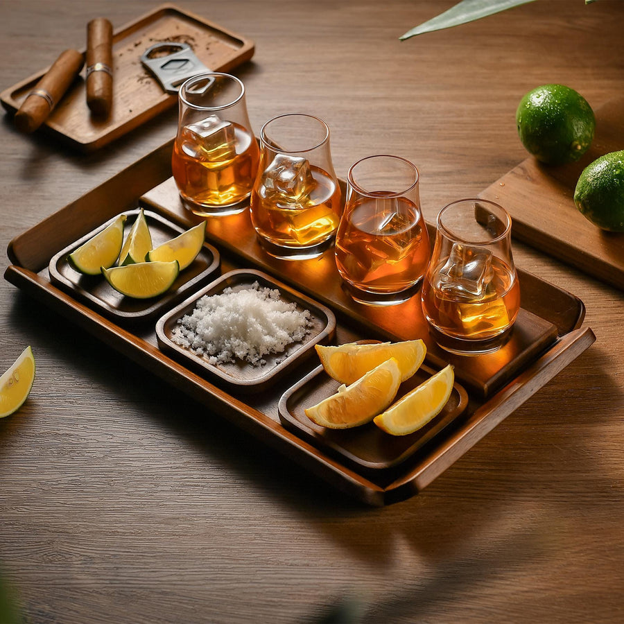 A tray with five glasses of amber-colored liquid, likely whiskey, and various garnishes such as lemon slices and salt. The tray is placed on a wooden surface, and there are also some limes and a wooden object in the background.