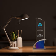 Academic Excellence Award for John Smith, 2024, displayed on a wooden desk. The award features a sleek, clear design with a blue gradient base, illuminated by a desk lamp, beside office supplies and a potted plant.