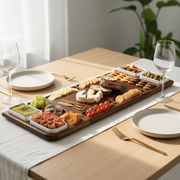 A wooden platter filled with an assortment of appetizers, cheeses, crackers, and other finger foods is displayed on a table, accompanied by two wine glasses and two forks.