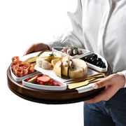 A person is holding a wooden tray with various food items, including meats, cheeses, bread, and fruits.