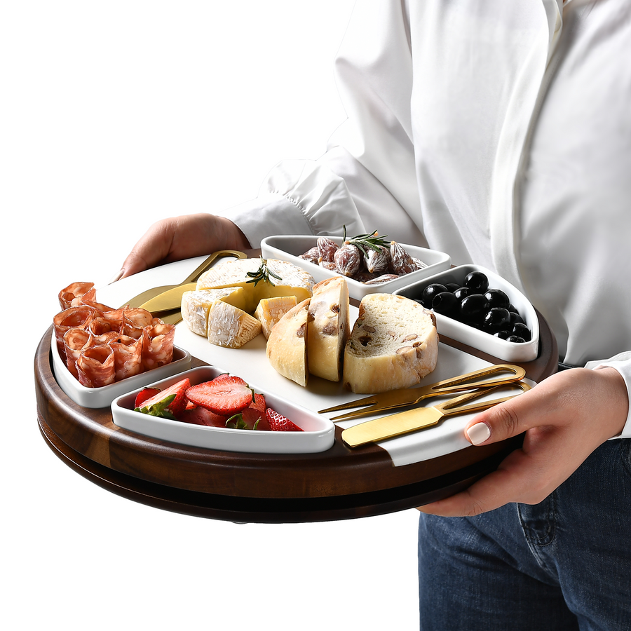 A person is holding a wooden tray with various food items, including meats, cheeses, bread, and fruits.